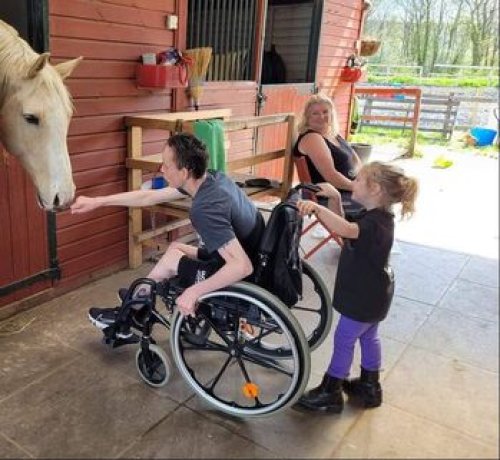 Photograph of a young man leaning froward from his wheelchair and allowing a white horse to smell his out reached fist. A much younger girl is holding the handles of the wheelchair . 