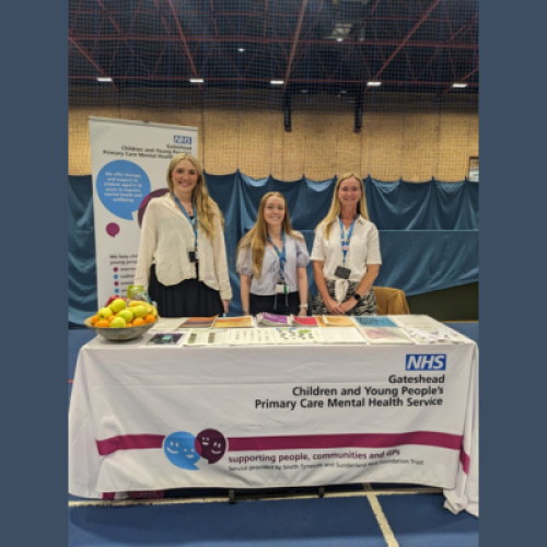 3 people stood behind a table promoting children's mental health services, which appears to be in a school gym