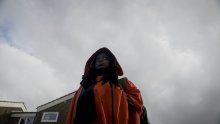 A girl stands in an orange raincoat with her hood up looking into the distance. The sky is grey filled with clouds in the background, the top part of a house is visible behind the figure. The image is a still from Larry Achiampong's feature length single channel 4K film, WayFinder (2022)