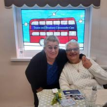 two people with an arm round each other smiling, in front of a sign for Teams and Bensham Community Care