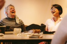 Two women laughing, sitting at a table with tea and biscuits.