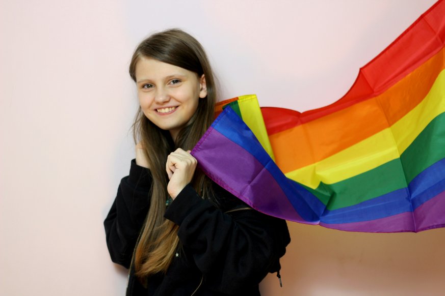 A smiling young person with the Pride flag over thier shoulders