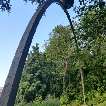 A steel, arched sculpture that is placed on a bank. The sculpture also has a steel ball attached to it on the top.