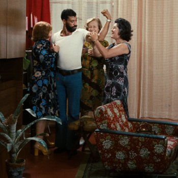 A man stands flexing his arm muscle whilst surrounded by a group of woman. 