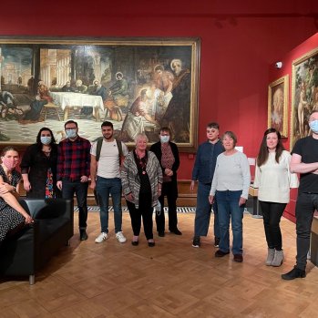 A group of people standing in line at the Shipley Art Gallery. Behind them is the large painting by Tintoretto.