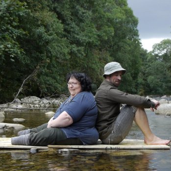 Julie Cleves and Robbie Synge sit back to back on wooden panels in a shallow stream. The background is filled with leafy green trees. 