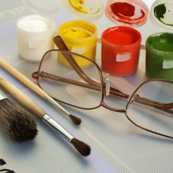 a photo of paint brushes and small paint pots surrounding a pair of discarded glasses 