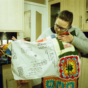colour photograph, interior. A Drag King with glasses and a knitted jumper looks down at his knitted blanket which he holds open across his chest. He stands in a kitchen.