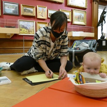 A photograph of a woman sitting on the floor in the Shipley Art Gallery drawing on yellow card. A baby is next to her on a playmat and lightly chewing a basket.