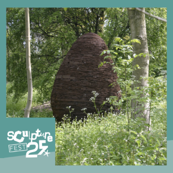 A photo that shows a pine cone sculpture standing in the middle of a forest. In the corner there is a logo saying 'Sculpture Fest 2025'.