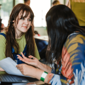 A teenage girl and an adult woman engaged in conversation across a table, with a laptop and notes between them at a mentoring event.